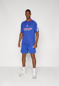 Sininen Adidas-urheiluasu, joka koostuu lyhythihaisesta paidasta punaisilla yksityiskohdilla ja logolla, yhdistettynä verkkomaisiin shortseihin, joissa on merkin logo.