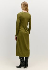 Langärmliges olivgrünes Kleid aus weichem Stoff, mit rundem Halsausschnitt und gerader Silhouette, kombiniert mit kniehohen schwarzen Stiefeln.