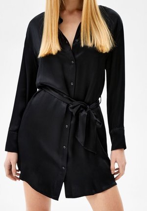 Woman wearing a black long-sleeve button-up dress with a tied waist belt and straight blonde hair, standing against a plain background.