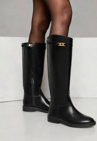 Botas de cuero negras hasta la rodilla con un acabado suave, que cuentan con un cierre lateral, una correa de acento dorado en la parte superior y una suela plana.