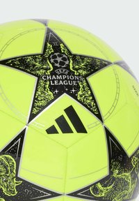 Žlutý fotbalový míč s černými prvky má hvězdový vzor, logo Ligy mistrů UEFA a značku Adidas. Hladká textura.