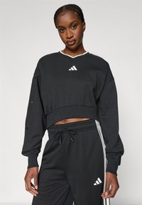 Μαύρο cropped φούτερ με λευκές λεπτομέρειες και εμβληματικό λογότυπο, συνδυασμένο με μαύρο αθλητικό παντελόνι από διχτυωτό ύφασμα με πλαϊνές ρίγες και ζώνη μέσης με κορδόνι.