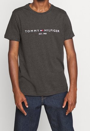 Mężczyzna w ciemnoszarej koszulce Tommy Hilfiger z logo i napisem „EST. 1985”, w zestawieniu z ciemnoniebieskimi dżinsami, stojący na jednolitym tle.