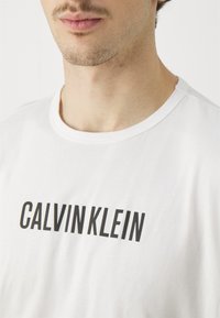 Biała bawełniana koszulka z czarnym logo "CALVIN KLEIN" w wyrazistych dużych literach, z okrągłym dekoltem i krótkimi rękawami.