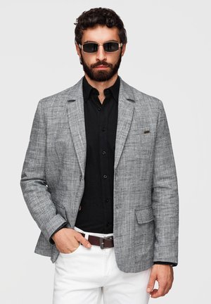 Homme barbu portant des lunettes de soleil, un blazer gris texturé, une chemise noire boutonnée, un pantalon blanc et une ceinture marron, debout avec une main dans la poche.