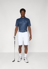 Polo marine à motif en losanges, associé à un short blanc. Les accessoires comprennent un gant de golf et un club. Chaussures : baskets blanches avec des accents.