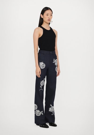 Donkerblauwe flared jeans met witte bloemenborduursels op de voorkant, gecombineerd met een aansluitend zwart tanktop en zwarte schoenen.