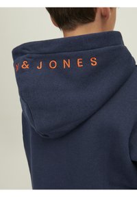 Námořnická modrá mikina s kapucí, vyrobená z měkké tkaniny, s oranžovým vyšitým logem na zádech. Má uvolněný střih a velkou kapuci.