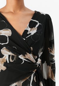 Robe portefeuille noire à motifs floraux beiges et blancs, avec un décolleté en V profond et un tissu délicat et texturé.