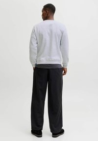 Man die met zijn rug naar de camera staat in een lichtgrijze sweatshirt, een donkergrijze t-shirt, losse zwarte broek, zwarte schoenen en een kleine ringoorbel, op een witte achtergrond.