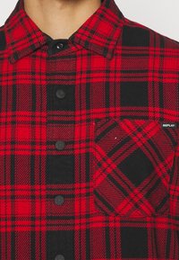 Camisa de franela a cuadros en rojo y negro con cierre de botones en el frente, bolsillos en contraste y una etiqueta de marca en el pecho. Diseño de tela texturizada.
