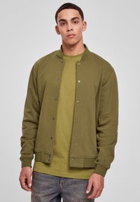 Ung mand med kort, mørkt hår iført en olivengrøn knap-op jakke over en matchende t-shirt og blå denim jeans, stående foran en ensfarvet grå baggrund.
