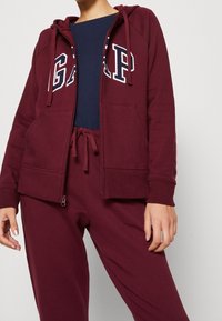 Felpa burgundy con zip e logo blu navy, tasche frontali, cappuccio con cordino e polsini a coste, abbinata a pantaloni coordinati. Materiale in morbido misto cotone.