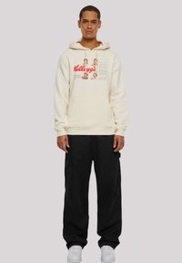 Cremefarbener Hoodie mit Fronttasche, versehen mit rotem "Kellogg's"-Grafik und Bildern. Kombiniert mit schwarzen, locker sitzenden Hosen und weißen Turnschuhen.