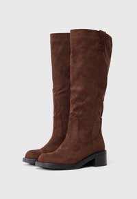 Botas de camurça castanhas até ao joelho com um salto robusto, apresentando costuras laterais e puxadores na parte superior para facilitar o uso. Textura suave e bico arredondado.