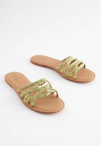 Platta sandaler med en ljusbrun, slät sula och flätade gröna och silverfärgade remmar. Öppen tå-design med flera korsande band för stöd.