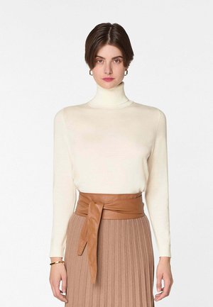 Femme aux cheveux bruns courts portant un pull à col roulé crème, des boucles d'oreilles créoles en or et une jupe plissée beige avec une large ceinture en cuir nouée à la taille.