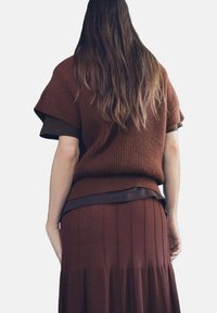 Suéter marrón de punto con mangas cortas, superpuesto sobre una blusa marrón oscura. El conjunto cuenta con una falda marrón plisada. Apariencia texturizada y acogedora.