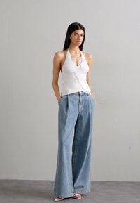 White ribbed halter top with lace trim paired with loose, light blue wide-leg jeans. Model stands with hands in pockets on a plain background.