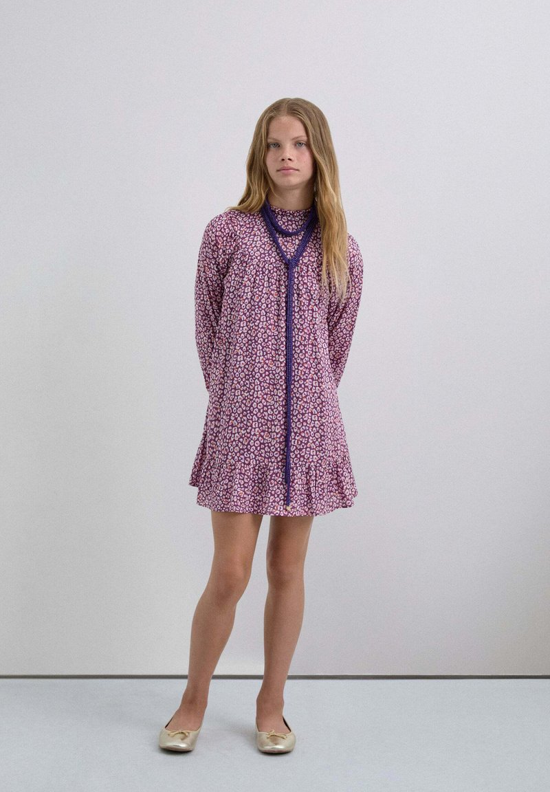Vestido de patrón morado y rosa con un dobladillo acampanado, mangas largas y un detalle de lazo en el escote. Tejido suave; combinado con bailarinas doradas.