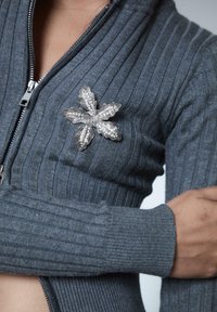 Osoba w szarym swetrze z zapięciem na zamek, o ribbowanym wzorze, z osadzonym przy klatce piersiowej srebrnym broszką w kształcie liścia z cyrkoniami.