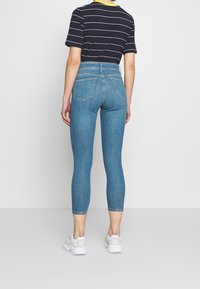 Osoba stojící zády, oblečená v krátkých modrých úzkých džínách, černobílém pruhovaném tričku s krátkým rukávem a bílých teniskách.