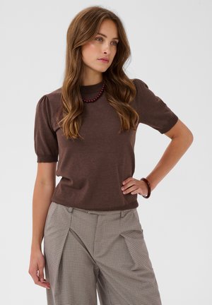 Femme aux longs cheveux bruns porte un haut marron à manches courtes, un pantalon large à carreaux, un collier de perles rouges et un bracelet assorti.