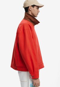 Rote Baumwolljacke mit braunem Kragen, die eine lockere Passform, seitliche Taschen und Knopfdetails aufweist, kombiniert mit einer beigen Kappe.