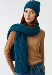 Ensemble bonnet et écharpe tricotés en bleu canard, pr�ésentant un motif texturé. L'écharpe se drape autour du cou, associée à un haut moulant marron.