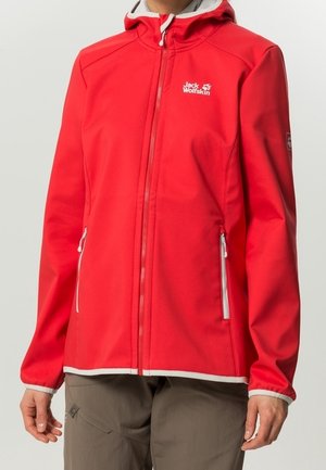 Veste rouge à capuche avec fermeture éclair frontale, poches latérales zippées et logo Jack Wolfskin sur la poitrine et la manche, portée avec un pantalon marron.