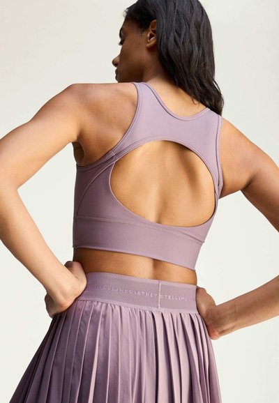 Woman with dark hair wearing a lavender sports bra with a back cutout and a matching pleated skirt, hands resting on her hips.