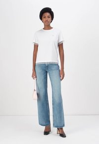 Camiseta blanca con un borde de encaje texturizado en las mangas, combinada con unos vaqueros de denim holgados. La modelo sostiene un pequeño bolso rosa y lleva tacones negros.