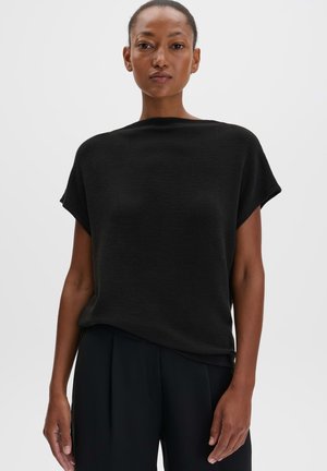 Femme aux cheveux courts portant un haut noir ample à manches courtes et un pantalon noir, debout devant un fond clair uni.