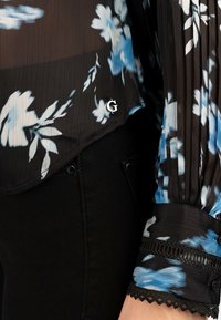 Blusa nera fluida con motivo floreale blu, tessuto plissettato e rifinitura in pizzo ai polsini. Presenta un piccolo logo argentato sull'orlo. Abbinata a pantaloni neri.