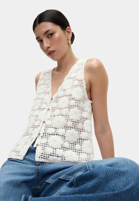 Weißer gehäkelter Weste mit Blumenmustern, V-Ausschnitt und Knöpfen vorne, ergänzt durch blaue High-Waist-Jeans.
