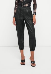 Pantalones cortos de cuero negro con un corte relajado, que cuentan con puños elásticos, múltiples bolsillos y una textura suave. Cierre de botón en la cintura.