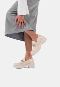 Scarpe platform in pelle verniciata beige con tacco chunky, caratterizzate da una cinghia decorativa e una superficie testurizzata, abbinate a una gonna di tessuto grigia.