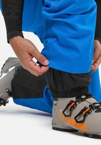 Blaue Schneehosen mit schwarzen Bündchen, elastischem Bund und einer Hand, die das Beinöffnung über grauen Skischuhen mit orangefarbenen Akzenten anpasst.