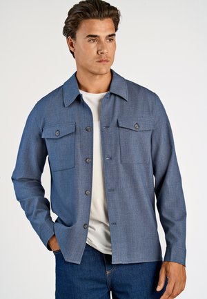 Jonge man die een blauw overhemd met borstzakken draagt over een wit T-shirt en donkerblauwe spijkerbroek, staande tegen een effen achtergrond.