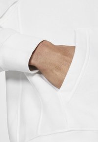 Un sweat à capuche blanc avec une poche kangourou. Texture douce visible, poignets côtelés et coutures détaillées sur les manches. Main insérée dans la poche.