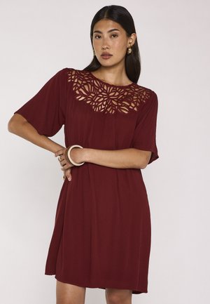 Donna che indossa un vestito bordeaux con un motivo floreale traforato sul petto, maniche corte e orecchini d'oro, posa con le braccia incrociate.