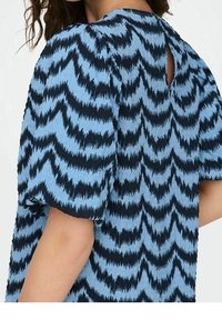 Blå kortärmad topp med en vågig svart mönstring, texturerad tyg och en nyckelhålsdetalj i ryggen vid halsringningen.