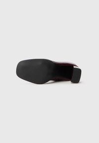 Una scarpa da uomo in pelle rosso scuro, sdraiata su un lato, che mostra la suola nera liscia e testurizzata e il tacco a blocco su sfondo bianco.