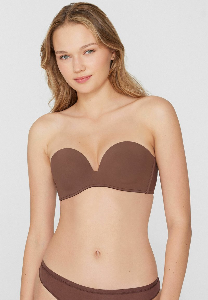 Junge Frau mit hellem Haar, die einen trägerlosen braunen BH und passende Unterwäsche trägt, vor einem schlichten hellen Hintergrund stehend.