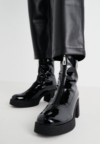 Botines de charol negro con un tacón de plataforma grueso, cremallera lateral y textura suave. Los botines se combinan con pantalones de cuero sintético negro.