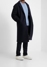 Cappotto di lana blu navy con colletto a punte, chiusura con bottoni e tasche laterali, indossato sopra una felpa azzurra, abbinato a jeans scuri e sneakers bianche.