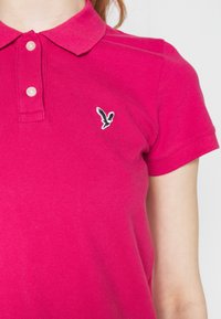Helles pinkes Poloshirt mit zwei Knöpfen und kleinem schwarz-weißen Vogel-Logo auf der linken Brust, getragen von einer Person mit heller Haut.