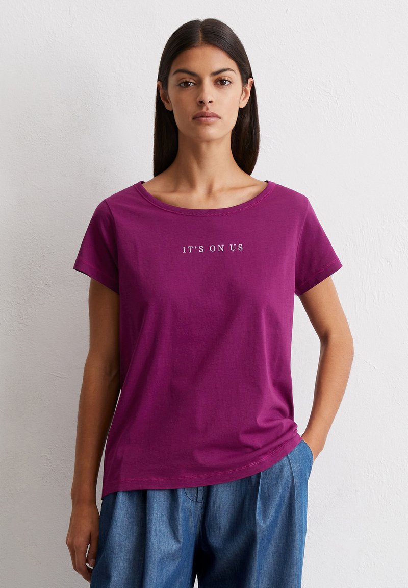 Lila T-Shirt mit kurzen Ärmeln und rundem Ausschnitt, bedruckt mit dem Text "WIR MACHEN DAS" in weiß. Aus weicher Baumwolle gefertigt, mit lockerer Passform und glatter Textur.