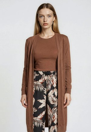 Ung kvinde med langt blondt hår iført en brun lang cardigan, matchende brun top og sorte bukser med store beige og brune bladmønstre.
