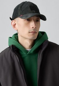 Gorra de corduroy negra con una superficie texturizada, detalle de parche negro y visera curva, combinada con una sudadera verde y una chaqueta oscura.